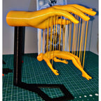 The Floating Marionette Illusion Sculpture | Desk & Home Decor (3D Printed) features a large hand holding strings, suspending a yellow figure mid-air. This unique yellow piece is displayed on a green grid cutting mat.